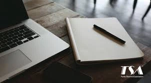 A laptop, a blank notebook, and a pen rest on a wooden table, with a smartphone partially visible.